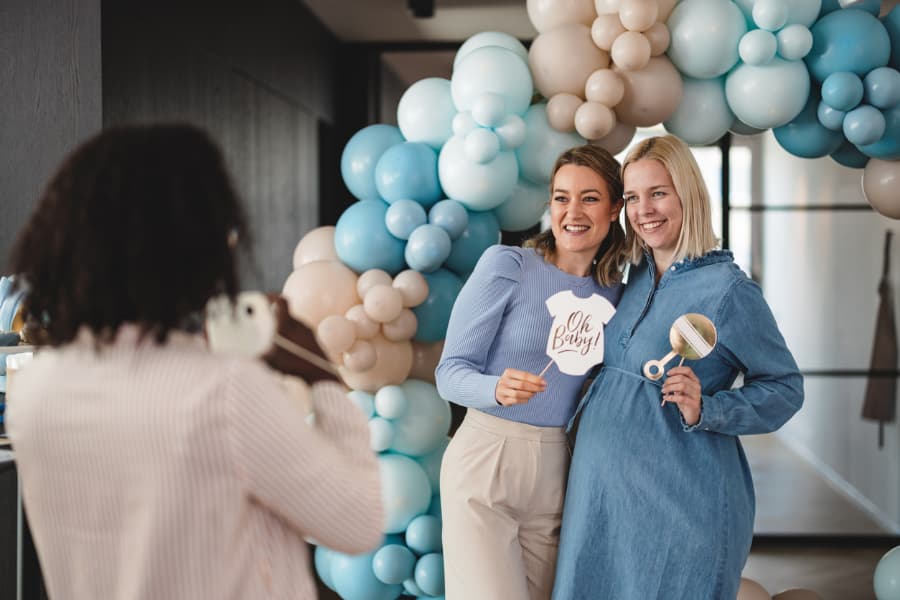 Two friends posing in front of balloon arch holding signs for baby shower
